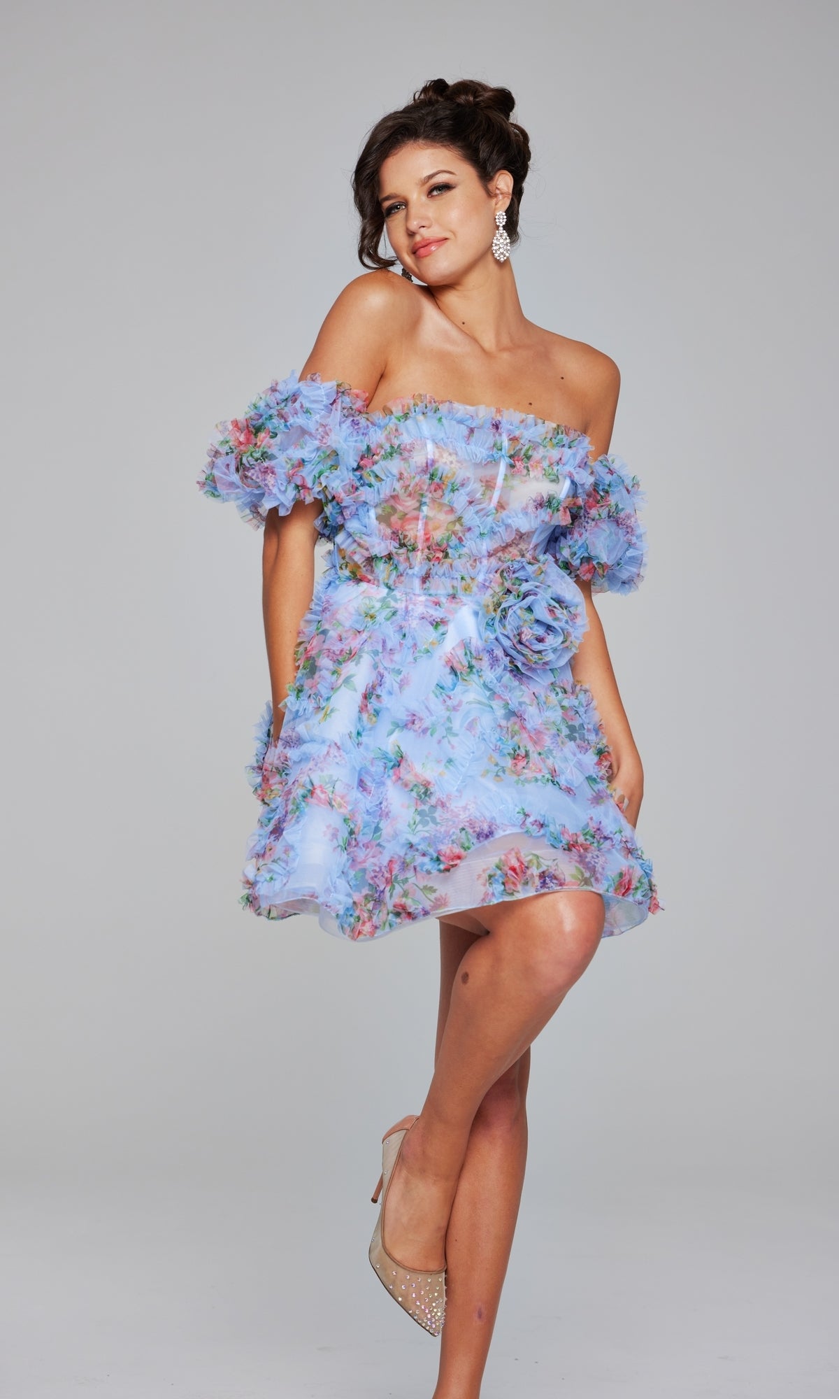 Woman modeling the Jovani Puff-Sleeve Short Print Hoco Dress 42573, featuring off-the-shoulder ruffled sleeves and bold floral print, paired with nude heels and statement earrings against a plain background.
